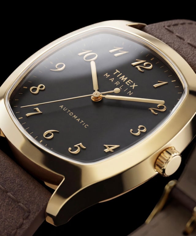 Крупный план Timex Marlin Chatham Automatic в золотистом корпусе с чёрным циферблатом, накладными золотистыми арабскими цифрами, золотистыми стрелками и коричневым кожаным ремешком