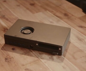 Компактная чёрная игровая консоль со встроенным дисководом, USB-портами и вентилятором охлаждения, расположенным в верхней части, лежит на деревянном столе.