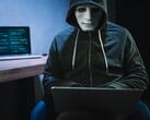 Фигура в маске и капюшоне работает на ноутбуке в затемненной комнате.