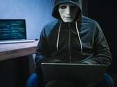 Фигура в маске и капюшоне работает на ноутбуке в затемненной комнате.