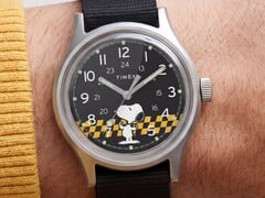Крупный план чёрного циферблата часов Timex MK1, на котором изображён Снупи на фоне жёлто-чёрной клетчатой графики с арабскими часовыми метками и светящимися стрелками.