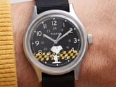 Крупный план чёрного циферблата часов Timex MK1, на котором изображён Снупи на фоне жёлто-чёрной клетчатой графики с арабскими часовыми метками и светящимися стрелками.
