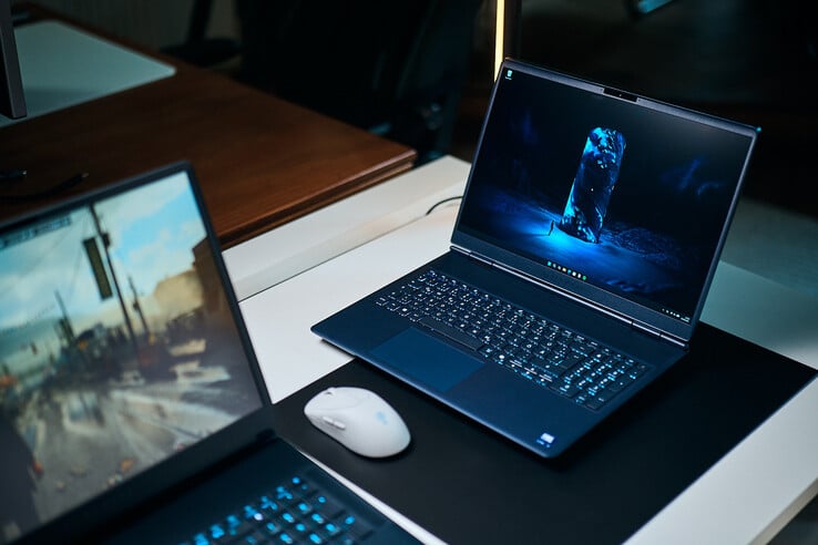 Тёмно-синий игровой ноутбук среднего класса (Alienware Aurora 16X), расположенный под углом на коврике для стола с сияющими синими обоями в стиле научной фантастики, стоит рядом с белой беспроводной мышью, а на переднем плане размыто виден ещё один ноутбук.