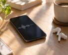 Смартфон с изображением звуковой волны на экране рядом с беспроводными наушниками и чашкой кофе на деревянном столе