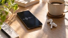 Смартфон с изображением звуковой волны на экране рядом с беспроводными наушниками и чашкой кофе на деревянном столе