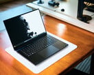 Игровой ноутбук Razer Blade 16 с открытым корпусом матового чёрного цвета, расположенный на белом коврике для стола, лежащем на тёплом деревянном столе, с яркими чёрно-белыми обоями на экране.