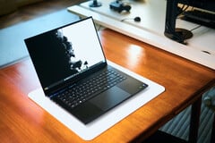 Игровой ноутбук Razer Blade 16 с открытым корпусом матового чёрного цвета, расположенный на белом коврике для стола, лежащем на тёплом деревянном столе, с яркими чёрно-белыми обоями на экране.