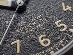 Крупный план текстурированного чёрного циферблата часов Timex Expedition Field Post Titanium с арабскими цифрами, светящимися стрелками и индексами, а также окошком даты.