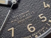 Крупный план текстурированного чёрного циферблата часов Timex Expedition Field Post Titanium с арабскими цифрами, светящимися стрелками и индексами, а также окошком даты.