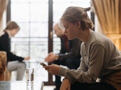 Девушка-подросток смотрит на смартфон, а на заднем плане вне фокуса сидят двое взрослых.