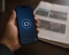 Рука держит смартфон с иконкой чат-бота, рядом лежит печатная газета.