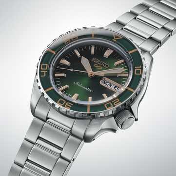 Крупный план Seiko 5 Sports SKX HDB005, демонстрирующий тёмно-зелёный циферблат, стрелки и накладные часовые метки цвета розового золота, а также соответствующий зелёный безель с разметкой золотистого цвета.
