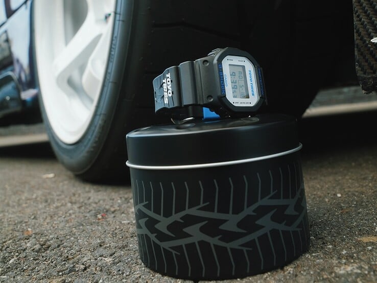 Часы Casio G-Shock DW5600TT25-1 x Toyo Tires, представленные на металлической коробке с изображением шины, расположенной рядом с автомобильным колесом.