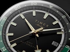 Крупный план чёрного циферблата Timex Atelier GMT Première M1a с золотистыми стрелками, люминесцентными часовыми метками, окошком даты и чёрно-зелёным 24-часовым кольцом GMT.