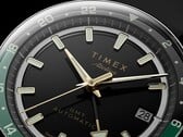 Крупный план чёрного циферблата Timex Atelier GMT Première M1a с золотистыми стрелками, люминесцентными часовыми метками, окошком даты и чёрно-зелёным 24-часовым кольцом GMT.
