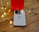 OnePlus 15 лежит на деревянном столе в окружении теплых гирлянд, а на заднем плане видна ярко-красная фирменная коробка OnePlus.