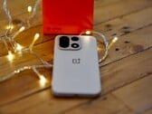 OnePlus 15 лежит на деревянном столе в окружении теплых гирлянд, а на заднем плане видна ярко-красная фирменная коробка OnePlus.