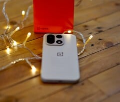 OnePlus 15 лежит на деревянном столе в окружении теплых гирлянд, а на заднем плане видна ярко-красная фирменная коробка OnePlus.