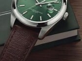 Крупный план часов Timex Legacy Day and Date: зелёный циферблат с отделкой «солнечные лучи», безель серебристого цвета и ушки с матовой шлифовкой, рифлёная заводная головка и фактурный коричневый кожаный ремешок на фоне тёмно-зелёной коробки и деревянной поверхности.