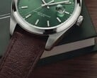 Крупный план часов Timex Legacy Day and Date: зелёный циферблат с отделкой «солнечные лучи», безель серебристого цвета и ушки с матовой шлифовкой, рифлёная заводная головка и фактурный коричневый кожаный ремешок на фоне тёмно-зелёной коробки и деревянной поверхности.