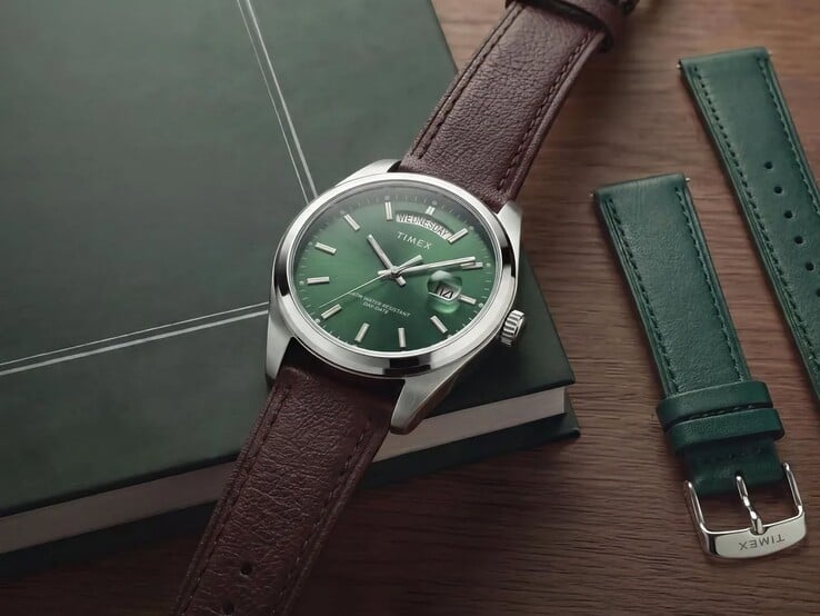 Timex Legacy Day and Date с зелёным циферблатом и коричневым кожаным ремешком на тёмно-зелёной книге рядом со сменным зелёным кожаным ремешком.