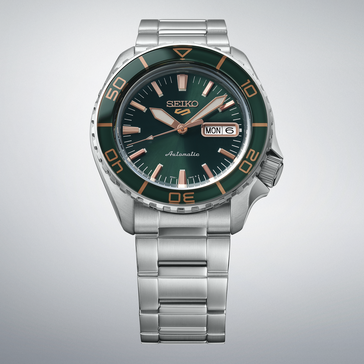 Seiko 5 Sports SKX HDB005 с темно-зеленым циферблатом, зеленым безелем в тон, стрелками и часовыми метками цвета розового золота и браслетом из нержавеющей стали на светло-сером фоне.