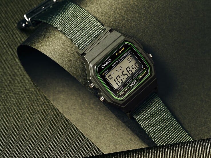 Цифровые часы Casio F-91WF-3A с чёрным корпусом с зелёными акцентами и плетеным ремешком из зелёной ткани, сфотографированные на тёмно-зелёном фактурном фоне.