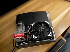 Изготовленная на заказ PlayStation 3 с водяным охлаждением, красным насосом SysCooling и большим черным вентилятором, установленным внутри корпуса, на деревянном столе. (Источник изображения: Zac Builds via YouTube)