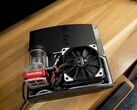Изготовленная на заказ PlayStation 3 с водяным охлаждением, красным насосом SysCooling и большим черным вентилятором, установленным внутри корпуса, на деревянном столе. (Источник изображения: Zac Builds via YouTube)