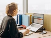 Женщина в очках работает с электронной таблицей на ноутбуке; на столе лежат папки и калькулятор