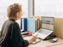 Женщина в очках работает с электронной таблицей на ноутбуке; на столе лежат папки и калькулятор