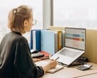 Женщина в очках работает с электронной таблицей на ноутбуке; на столе лежат папки и калькулятор