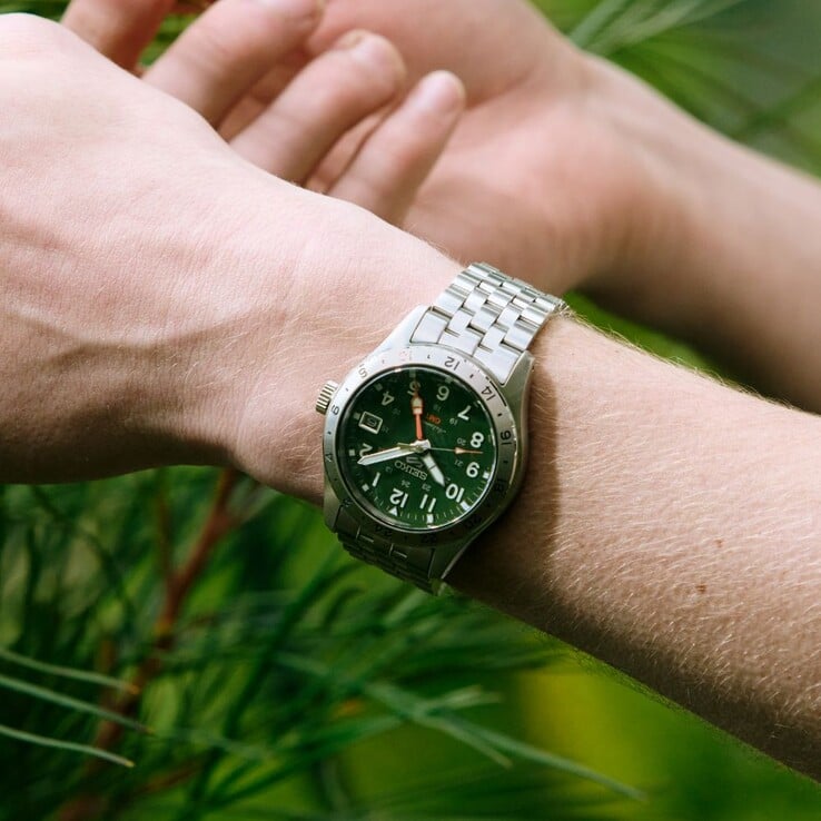 Вид спереди на часы Seiko 5 Sports Field GMT HDB001 с зелёным циферблатом, фиксированным 24-часовым безелем из нержавеющей стали, оранжевой стрелкой GMT и пятизвенным стальным браслетом.