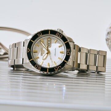 Часы Seiko 5 Sports SKX HDB004 с серебристо-белым циферблатом, черным вращающимся безелем с золотистыми отметками, золотистыми стрелками и индексами и браслетом из нержавеющей стали, снятые под углом.