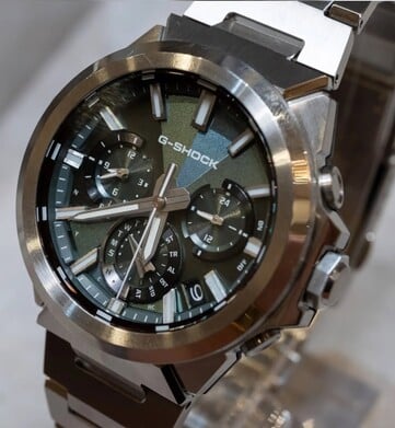 Крупный план часов Casio G-Shock GST-W1000D-3A с зелёным циферблатом, на котором видны его геометрическая текстура, три дополнительных циферблата, светящиеся метки и полированный безель из нержавеющей стали.