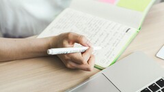 Человек делает записи в блокноте, сидя рядом с открытым ноутбуком