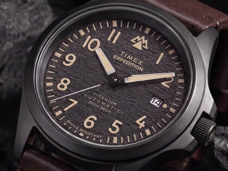 Крупный план текстурированного чёрного циферблата часов Timex Expedition Field Post Titanium с арабскими цифрами, светящимися стрелками и индексами, а также окошком даты.
