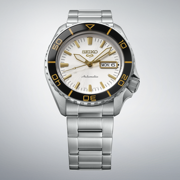 Seiko 5 Sports SKX HDB004 с серебристо-белым циферблатом, черным безелем с разметкой золотого цвета, стрелками и часовыми метками золотистого оттенка и браслетом из нержавеющей стали на светло-сером фоне.